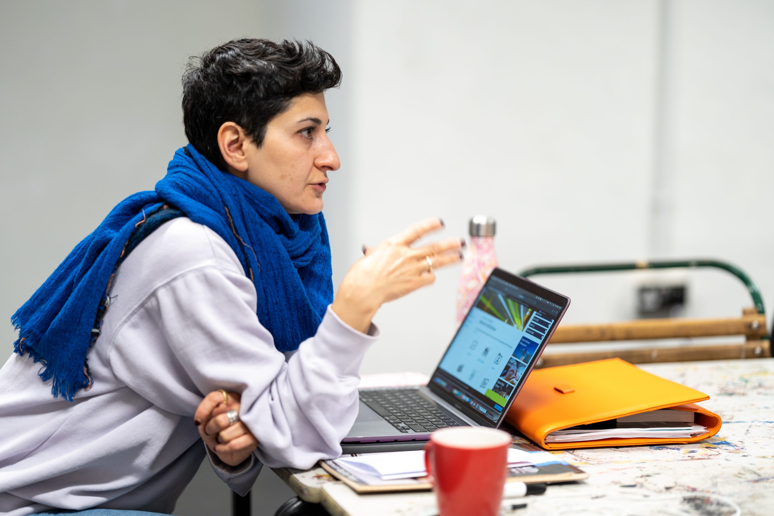 A lecturer talks while gesticulating. She has her computer and notebook with her. In the frame, we can see a messy, colourful table for art projects and a cup of coffee.