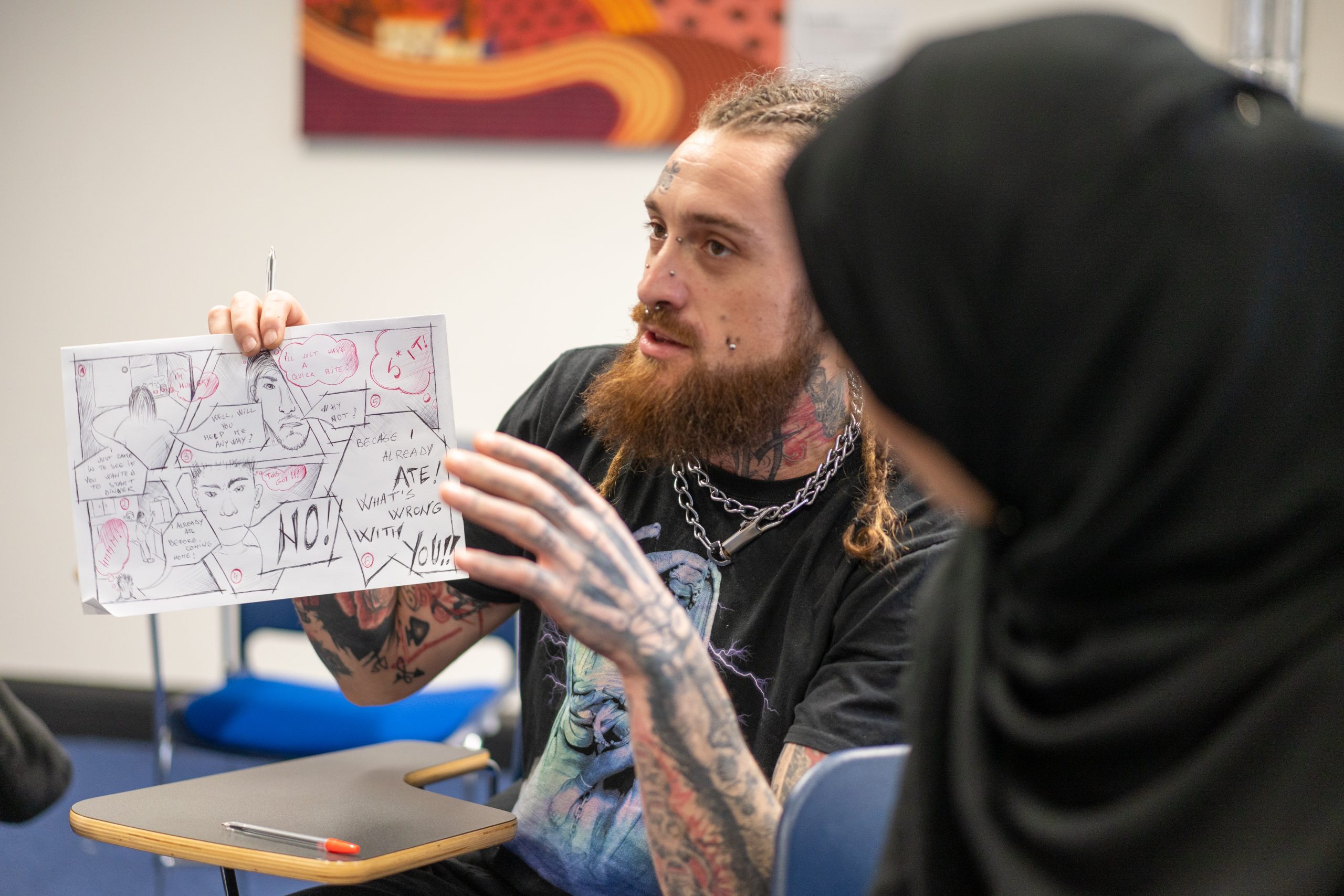 One student holds a draft of a new artwork project. He seems to be explaining what the project is about. Another student looks at him, listening carefully.