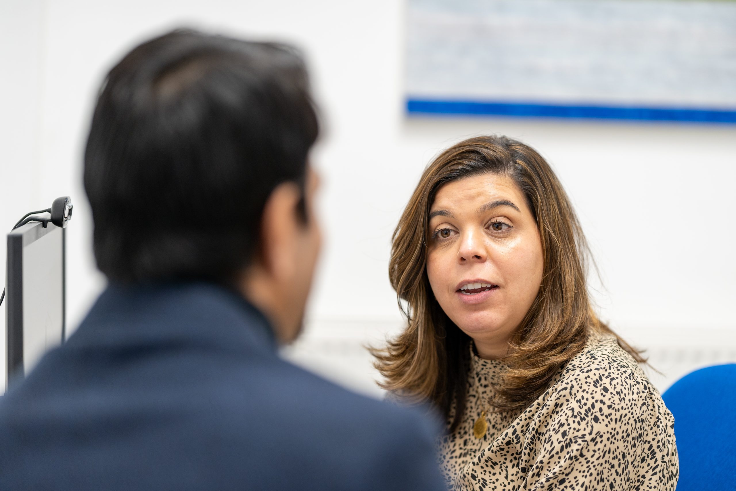 A woman is talking to a man in a blue chair. The woman faces the camera while the man has his back to the viewer. We can notice that the woman is helping the men with online documents.