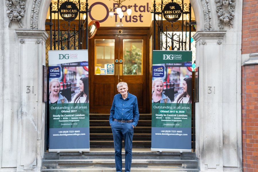 David Game poses in front of the DGHE Building with his hand in his pockets. You can see the college building and banners with information about courses.